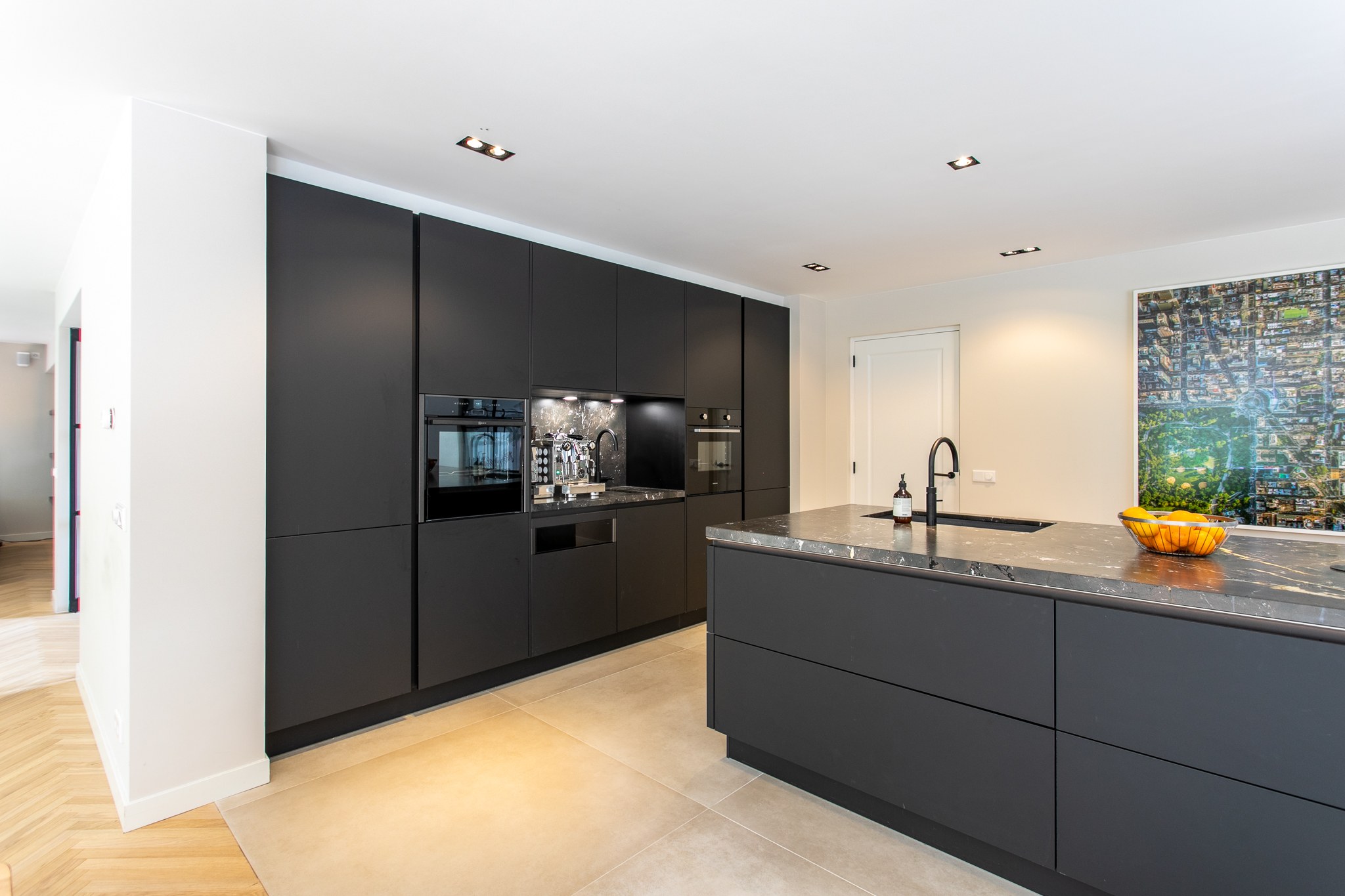 Moderne keuken in Blaricum met kamerhoge matzwarte kastenwand, zwart kookeiland, donker natuurstenen werkblad, lichte vloer en kleurrijk kunstwerk.