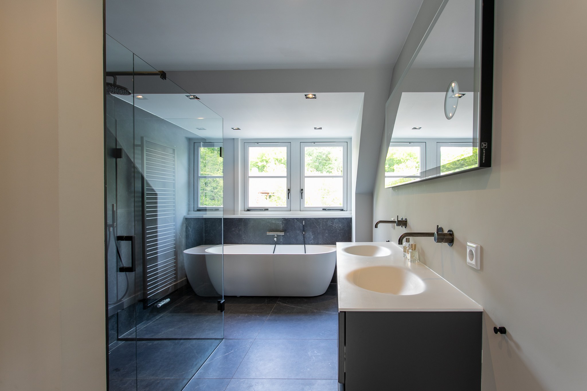 Strakke badkamer met groot vrijstaand wit bad, dubbele witte wastafel, donkergrijze tegelvloer, glazen inloopdouche en minimalistische grijze meubels.