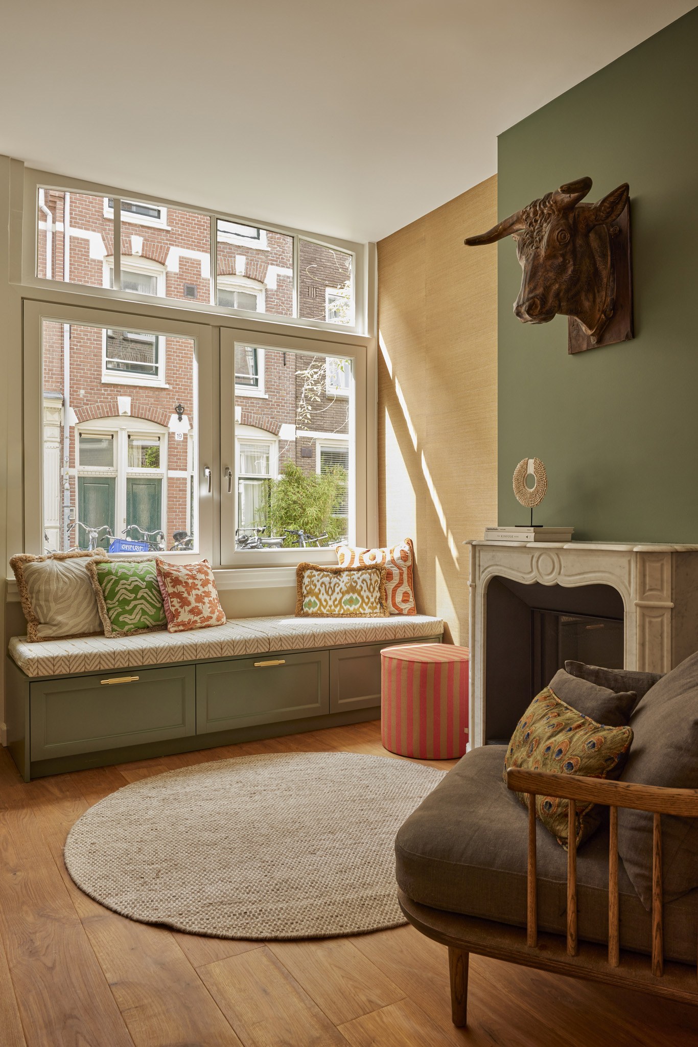 Warme woonkamer met olijfgroene wand, ingebouwde erkerbank, haard en visgraatvloer.