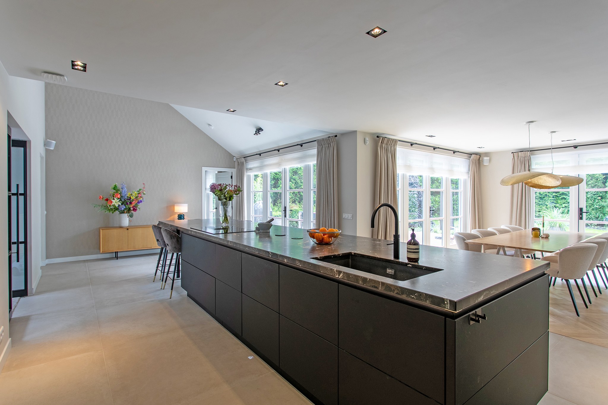 Lichte open leefkeuken in Blaricum met groot matzwart kookeiland, donker natuurstenen werkblad, eethoek, lichte gordijnen en zachte grijsbeige wanden.