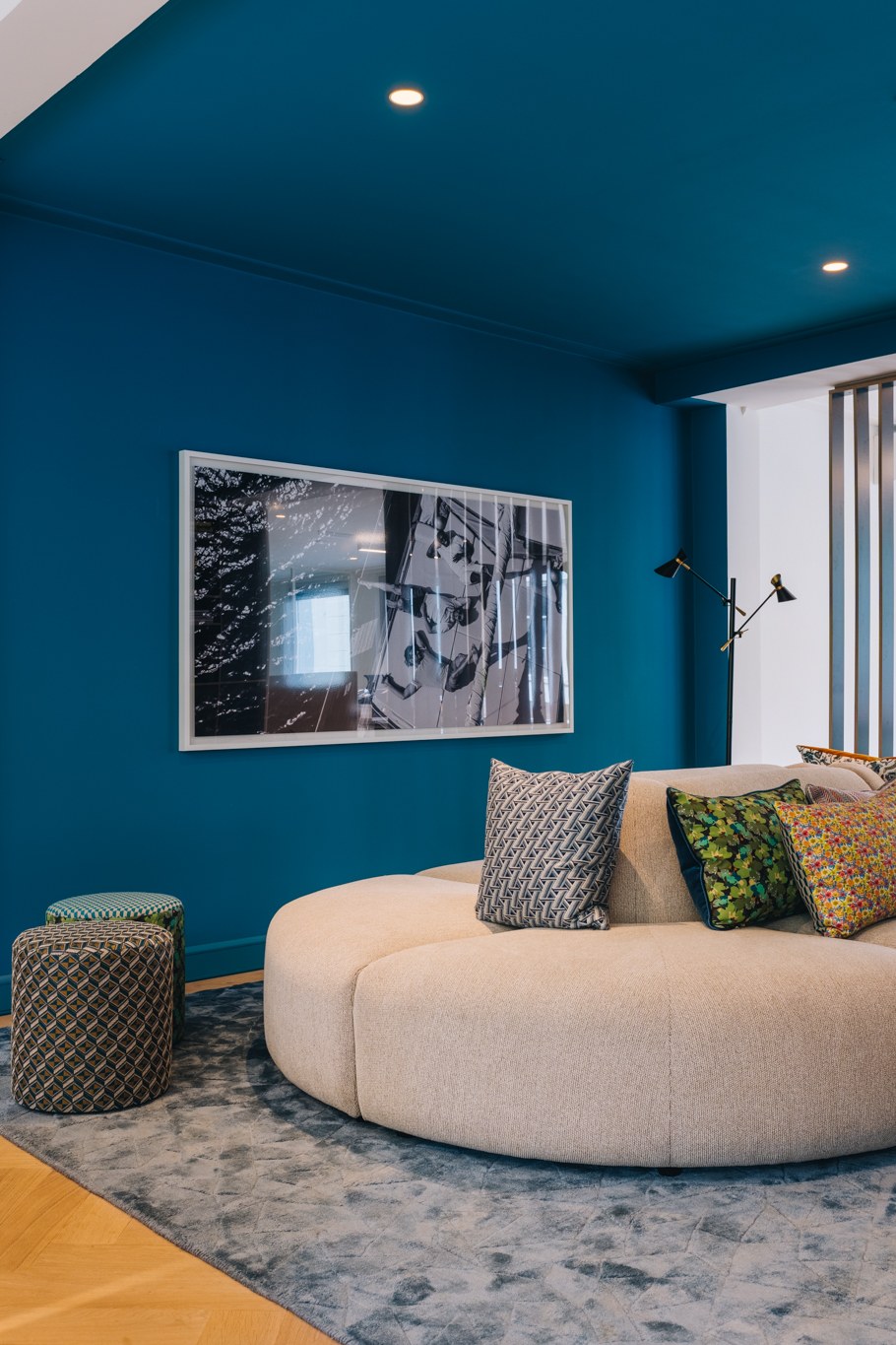 Verticale detailopname van een kantoorlounge met petrolblauwe wand, afgeronde crèmekleurige bank, kleurrijke kussens, patroonpoefs en kunstfotografie.