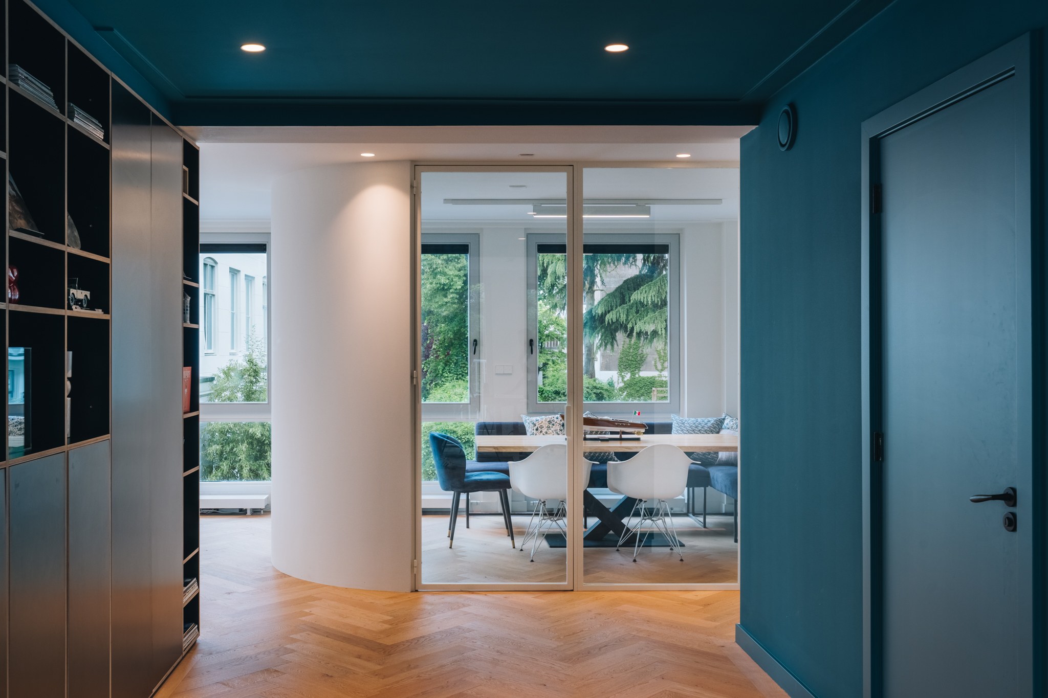 Doorkijk in een modern kantoor met diepe petrolblauwe wanden en plafond, zwarte maatwerkkast, visgraatvloer en glazen schuifwand naar de vergaderruimte.