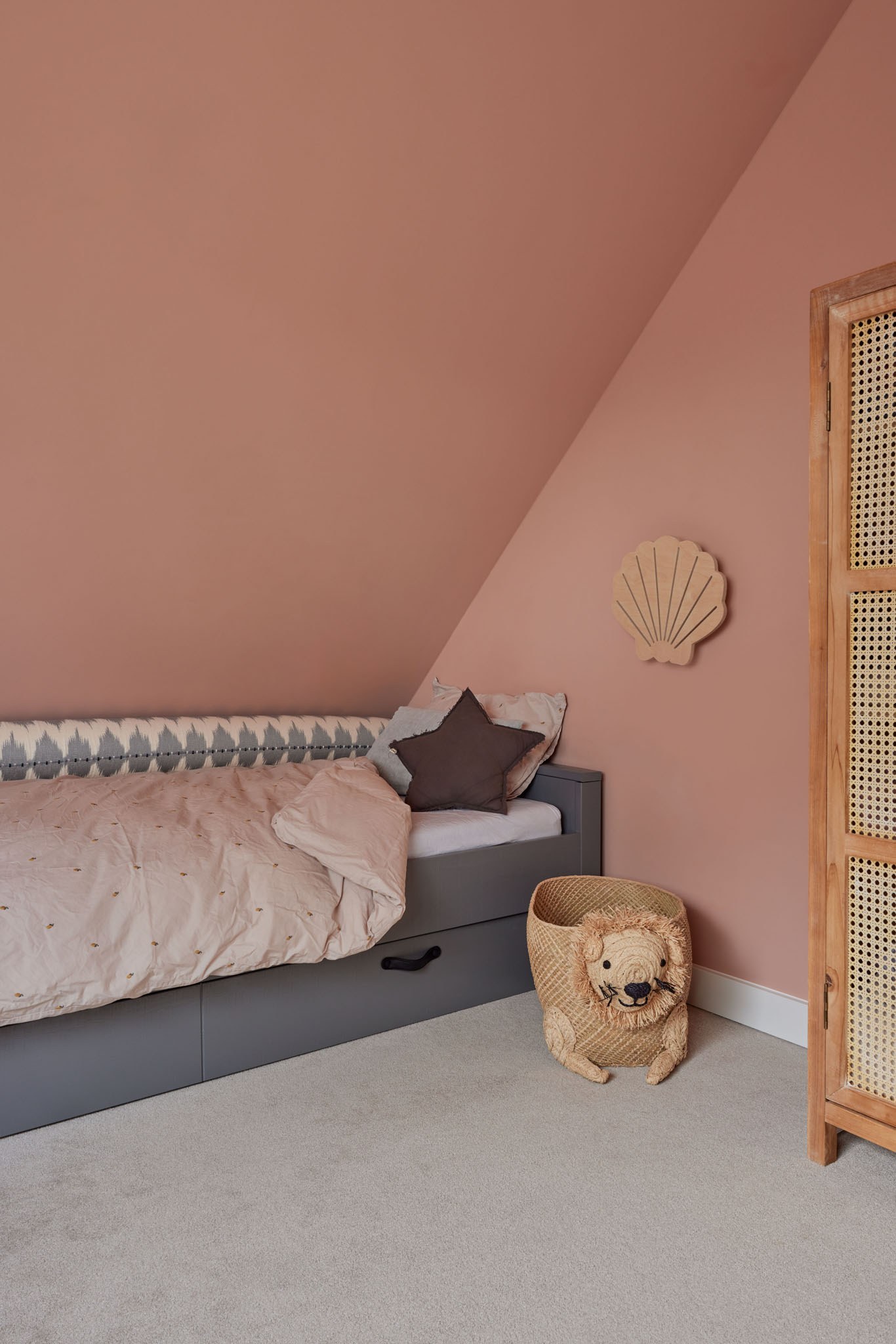 Zachtroze kinderkamer met grijze daybed, rotan kledingkast en warme textielaccenten.