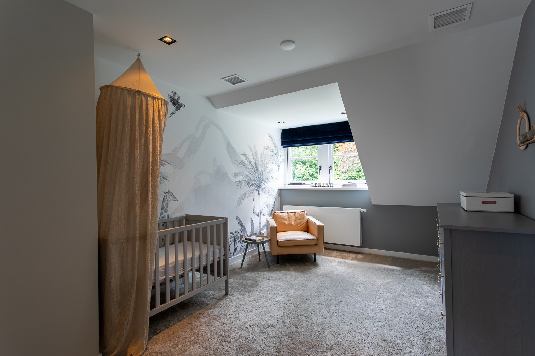 Lichte babykamer onder een schuin dak met grijze wanden, zachte wandillustratie, grijs ledikant, mosterdkleurige fauteuil en naturel bedhemel.