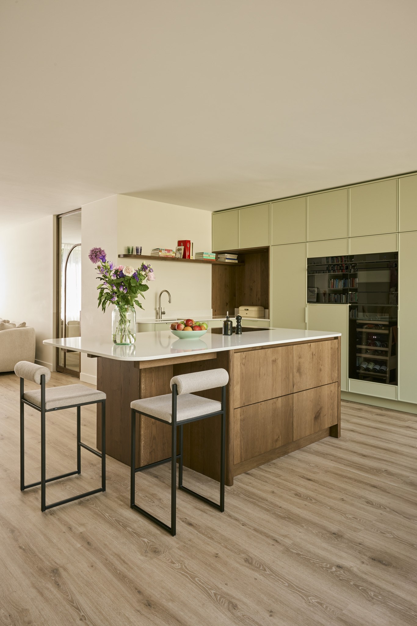 Open keuken met saliegroene maatwerkkasten, eiken kookeiland, licht werkblad, zwarte barkrukken en houtlook vloer.