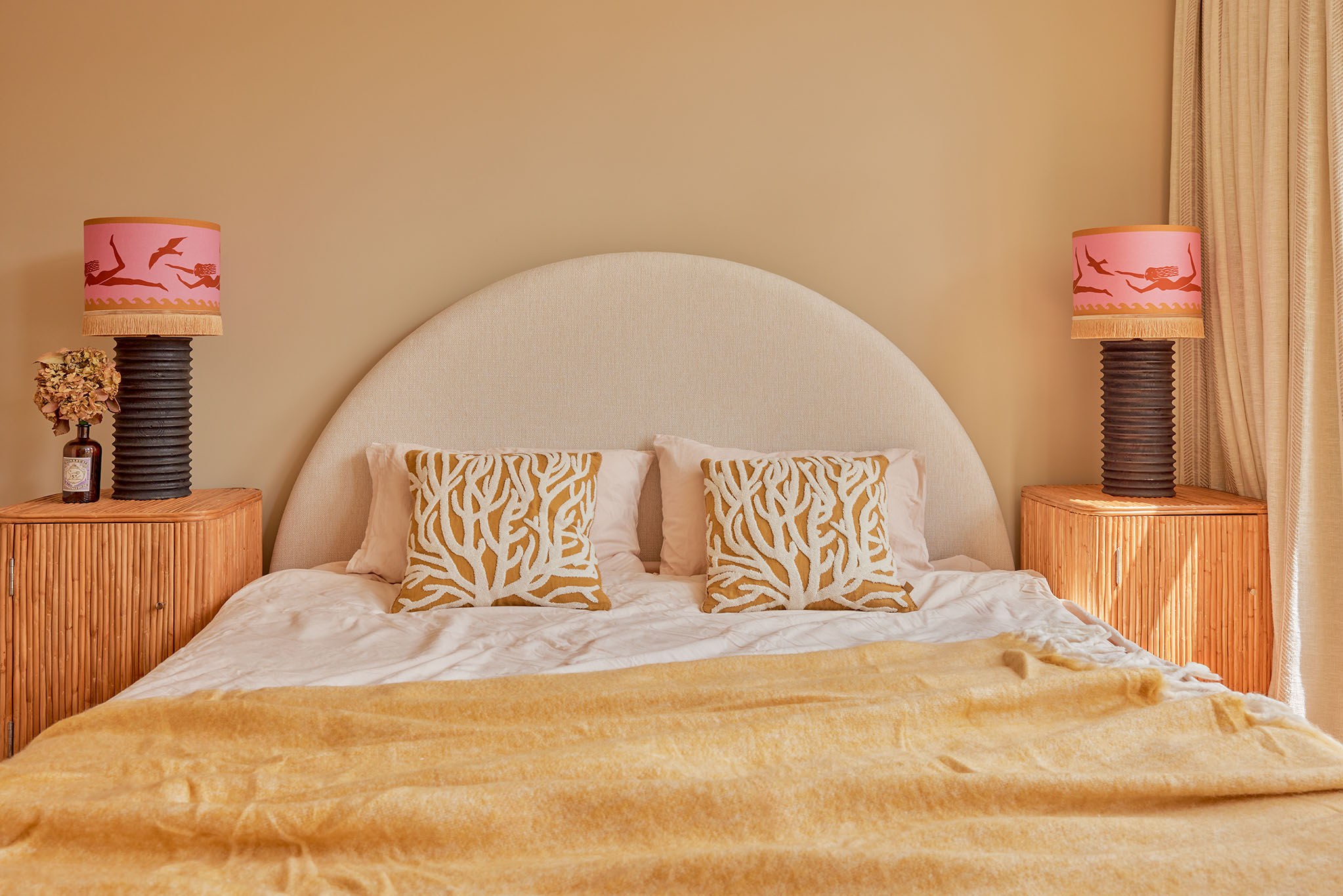 Slaapkamer met afgerond crèmekleurig hoofdbord, rotan nachtkasten, amberkleurige plaid en roze lampen.