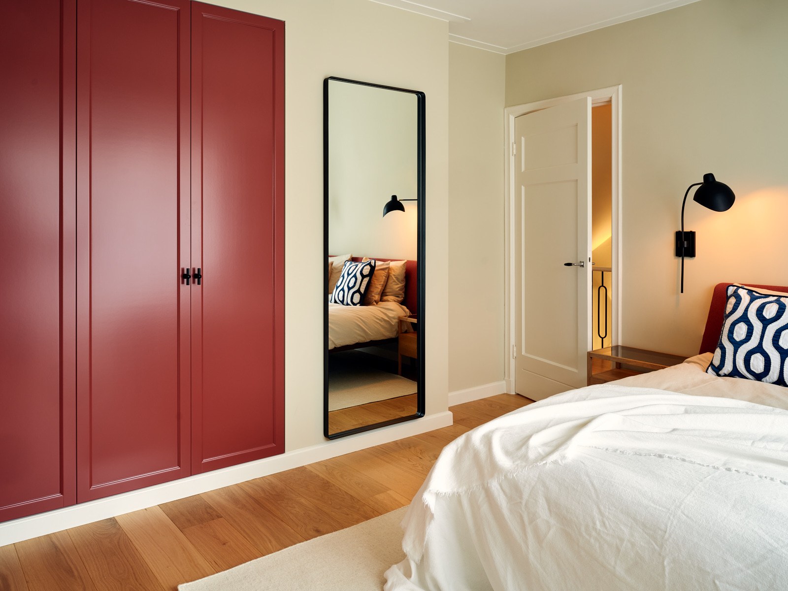 Warme slaapkamer in Bussum met diepe roestrode maatwerkkast, zwart omlijste spiegel, beige wanden, houten vloer en grafische blauw-witte textielaccenten.
