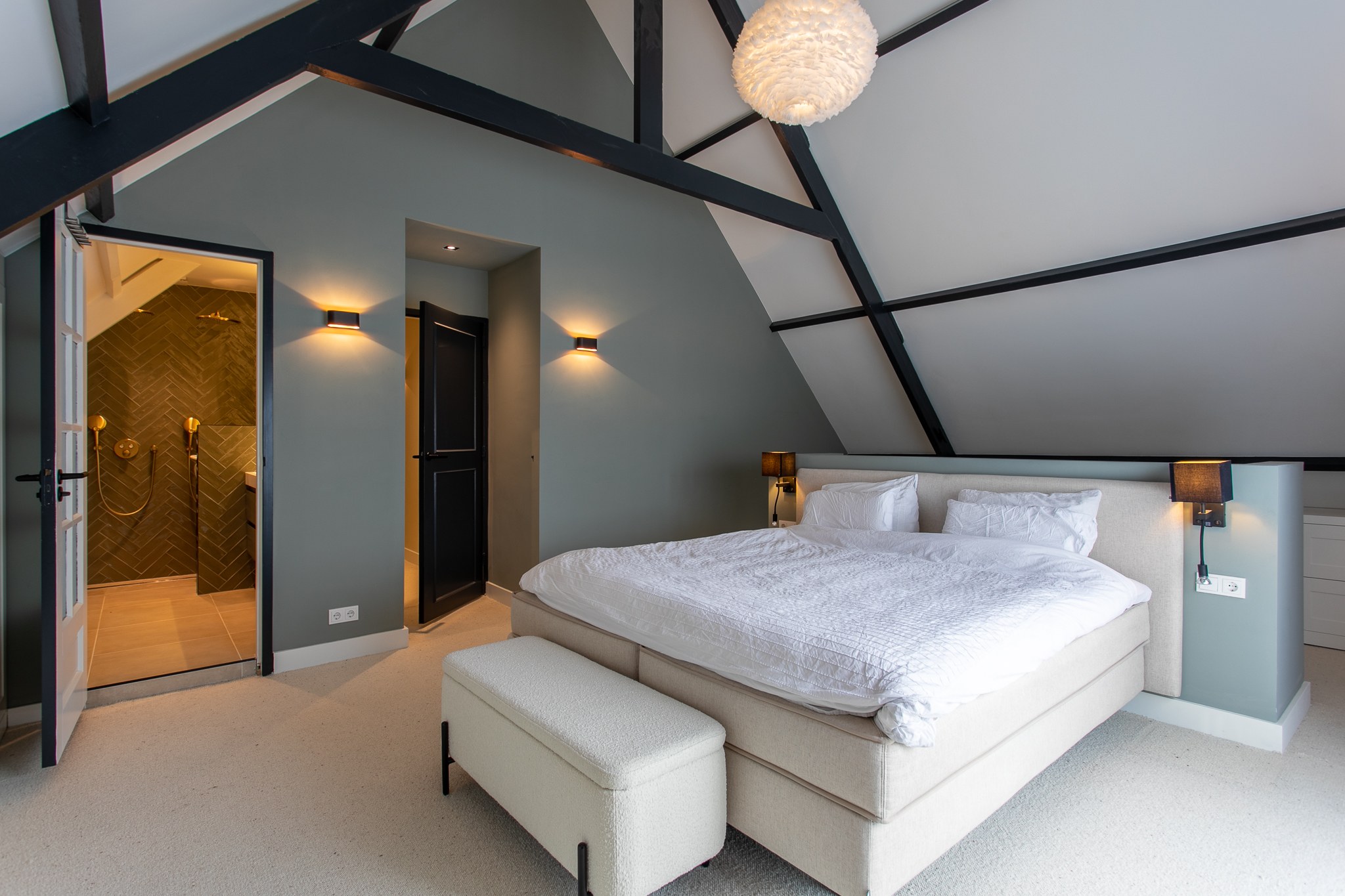 Ruime slaapkamer onder een schuin dak met zichtbare zwarte houten spanten, grijsgroene wanden, wit bed, zacht tapijt en warme wandverlichting.