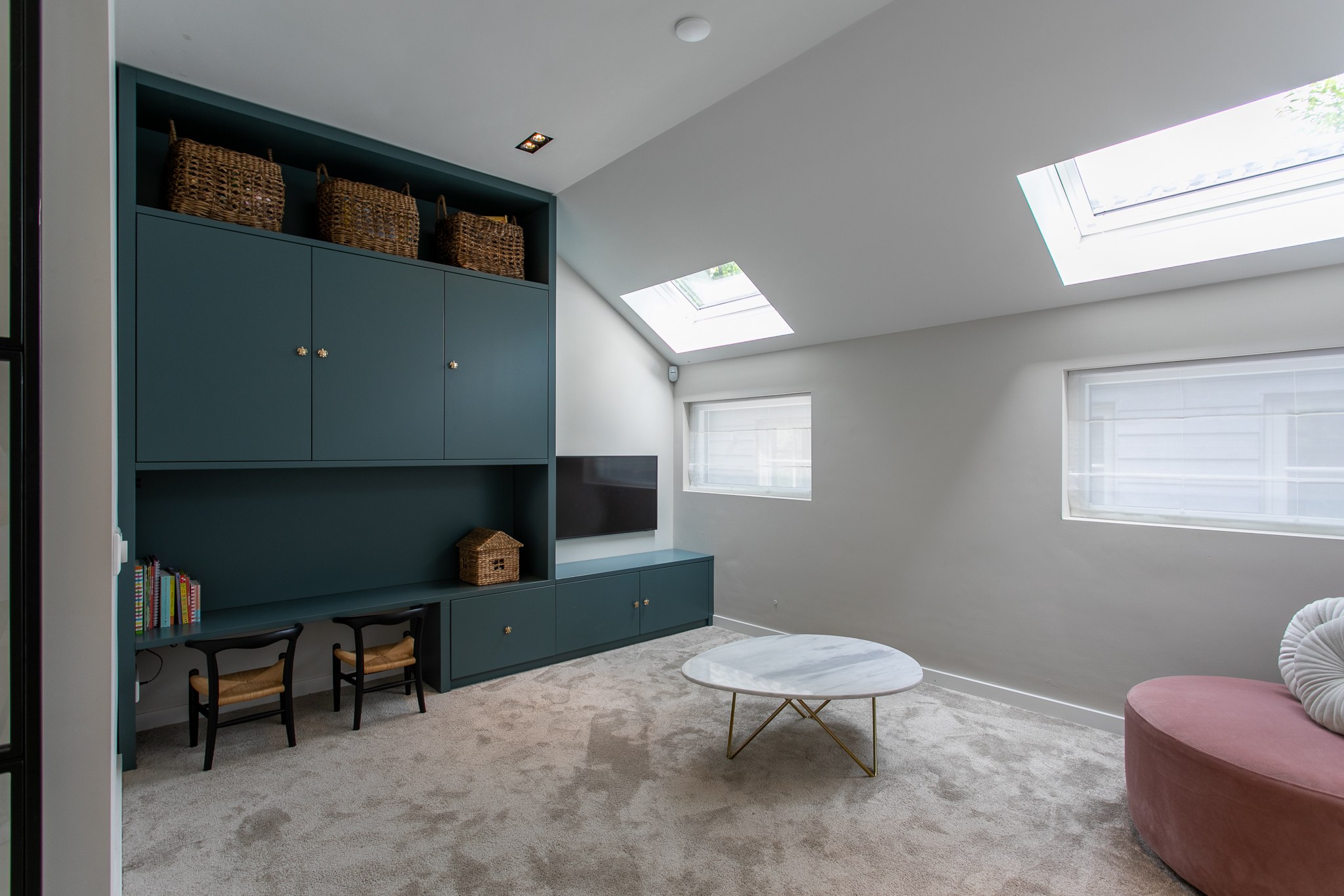 Speelse kinderkamer met petrolkleurige maatwerkkast, geïntegreerd speelhuis en bankje, zacht vloerkleed, lichtgrijze wand en poederroze ronde bank.