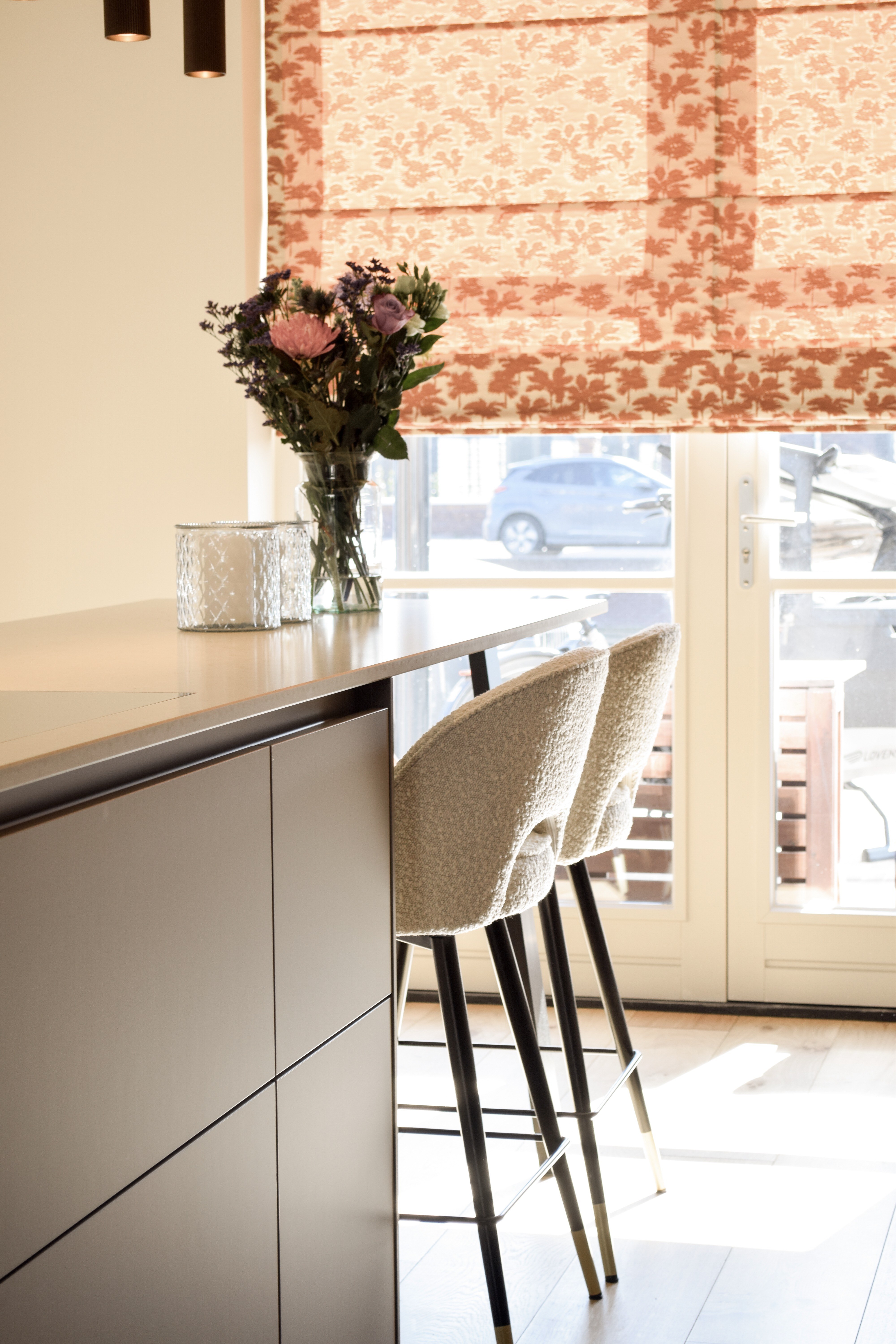 Taupe maatwerkkeuken met kookeiland, barstoelen en rood gebloemd vouwgordijn.