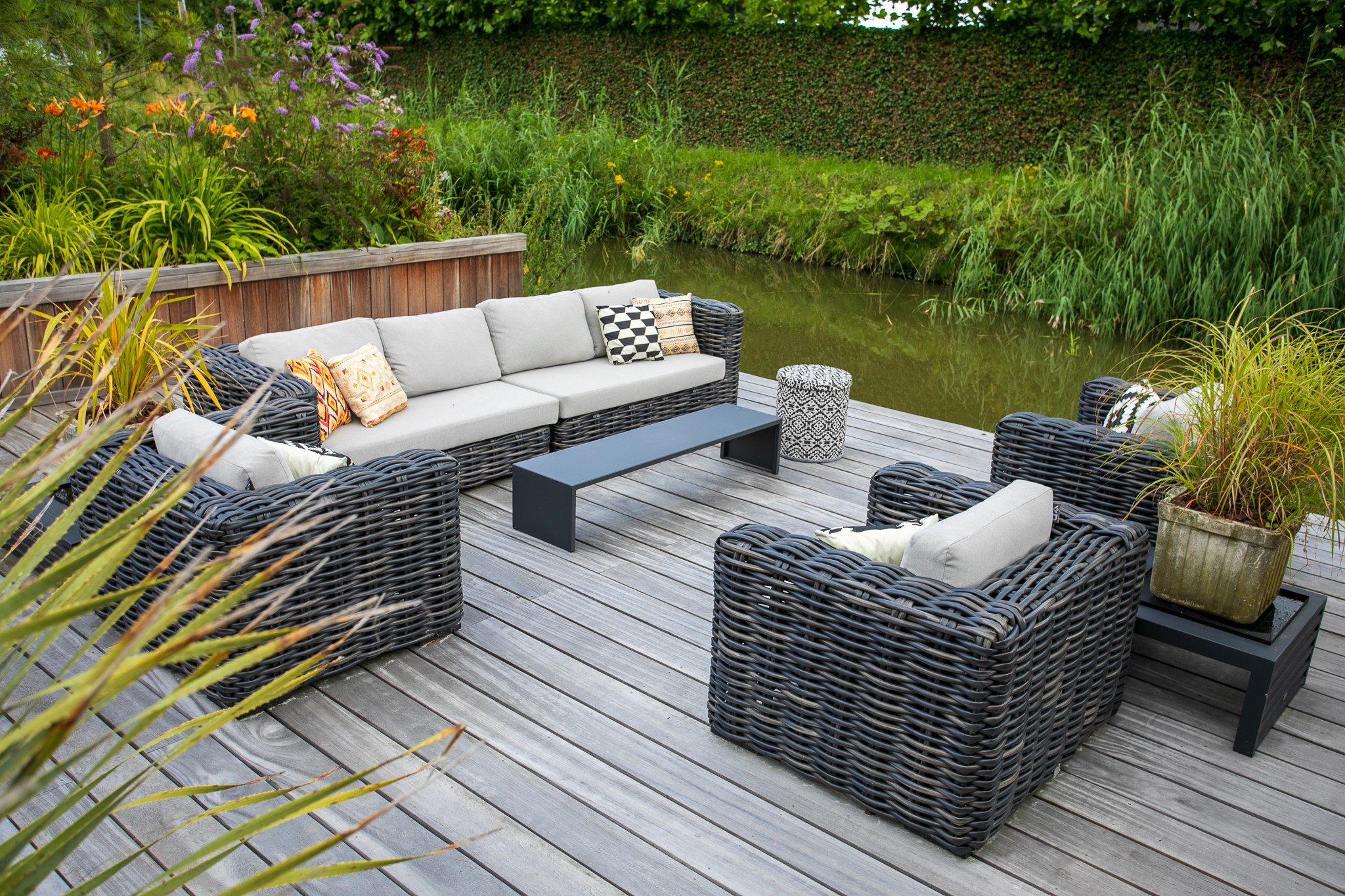 Sfeervol houten terras aan het water met grijze gevlochten loungeset, lichte kussens, zwarte salontafel en weelderige groene beplanting.