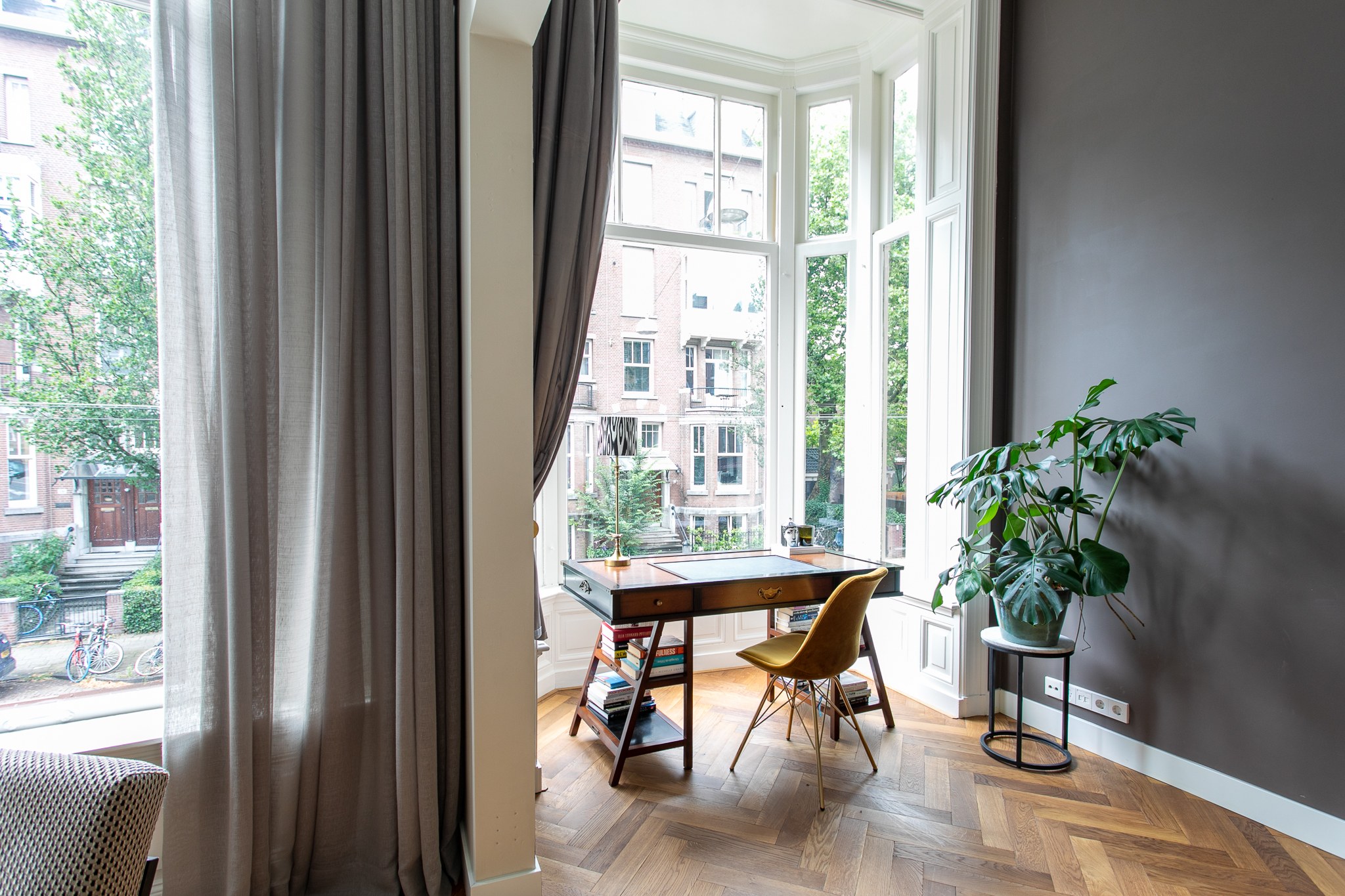 Licht thuiskantoor aan een hoge erker met klassieke ramen, donker houten bureau, okergele stoel, grote groene plant, grijze wand en visgraatvloer.