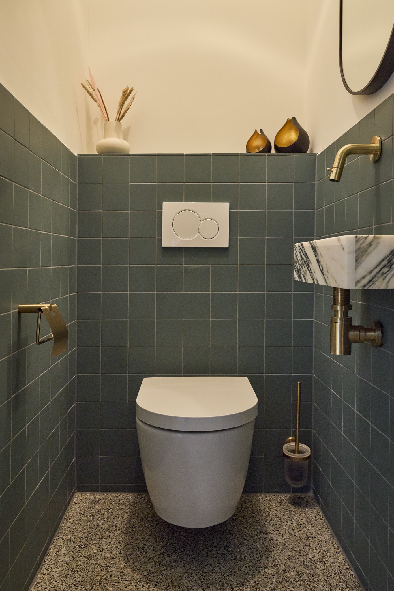 Toiletruimte met blauwgroene vierkante tegels, terrazzovloer, messing kranen en marmeren fontein.