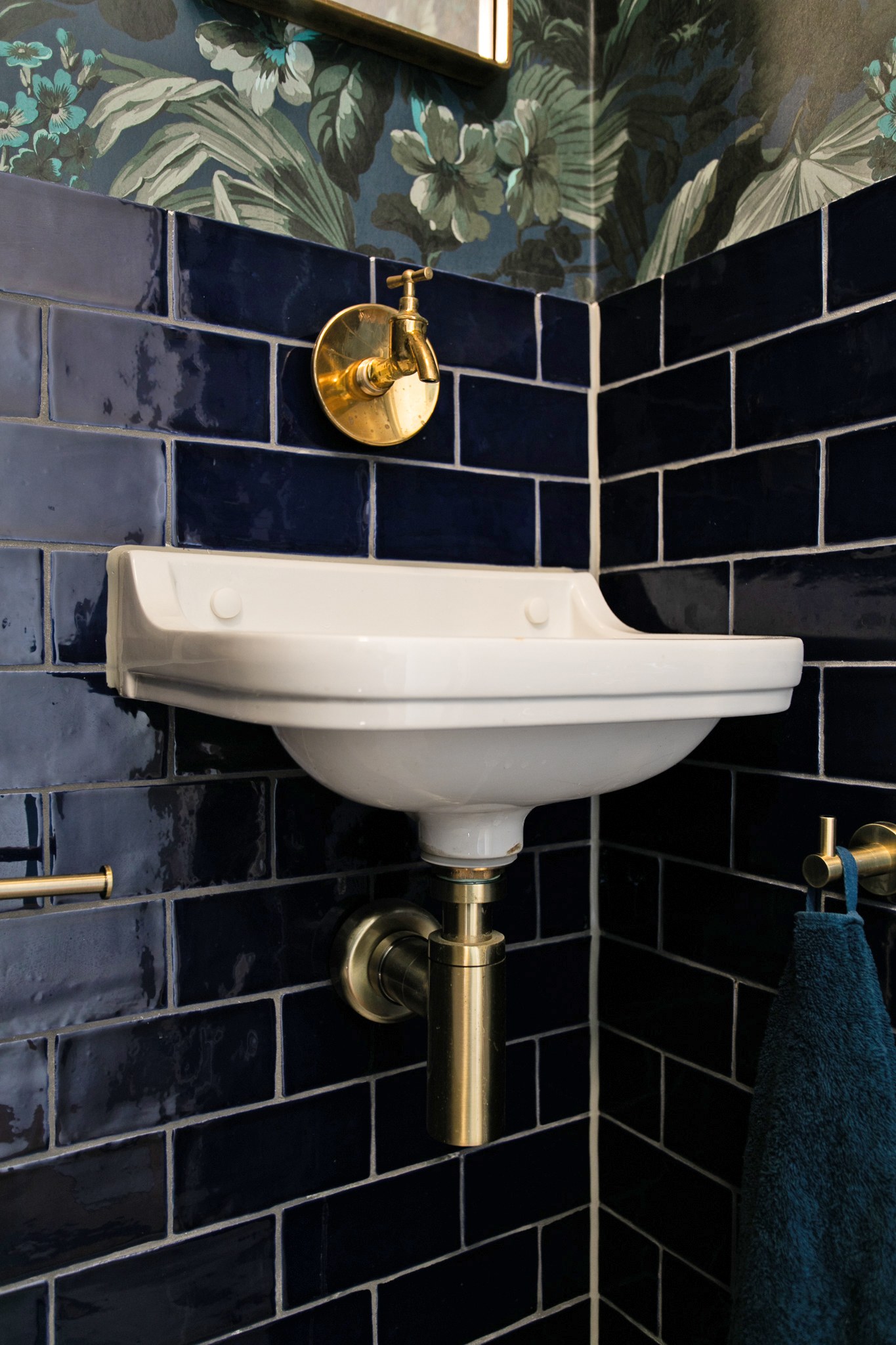Kleine toiletruimte met glanzende donkerblauwe metrotegels, botanisch behang, wit klassiek fonteintje en messing kraan en sifon.