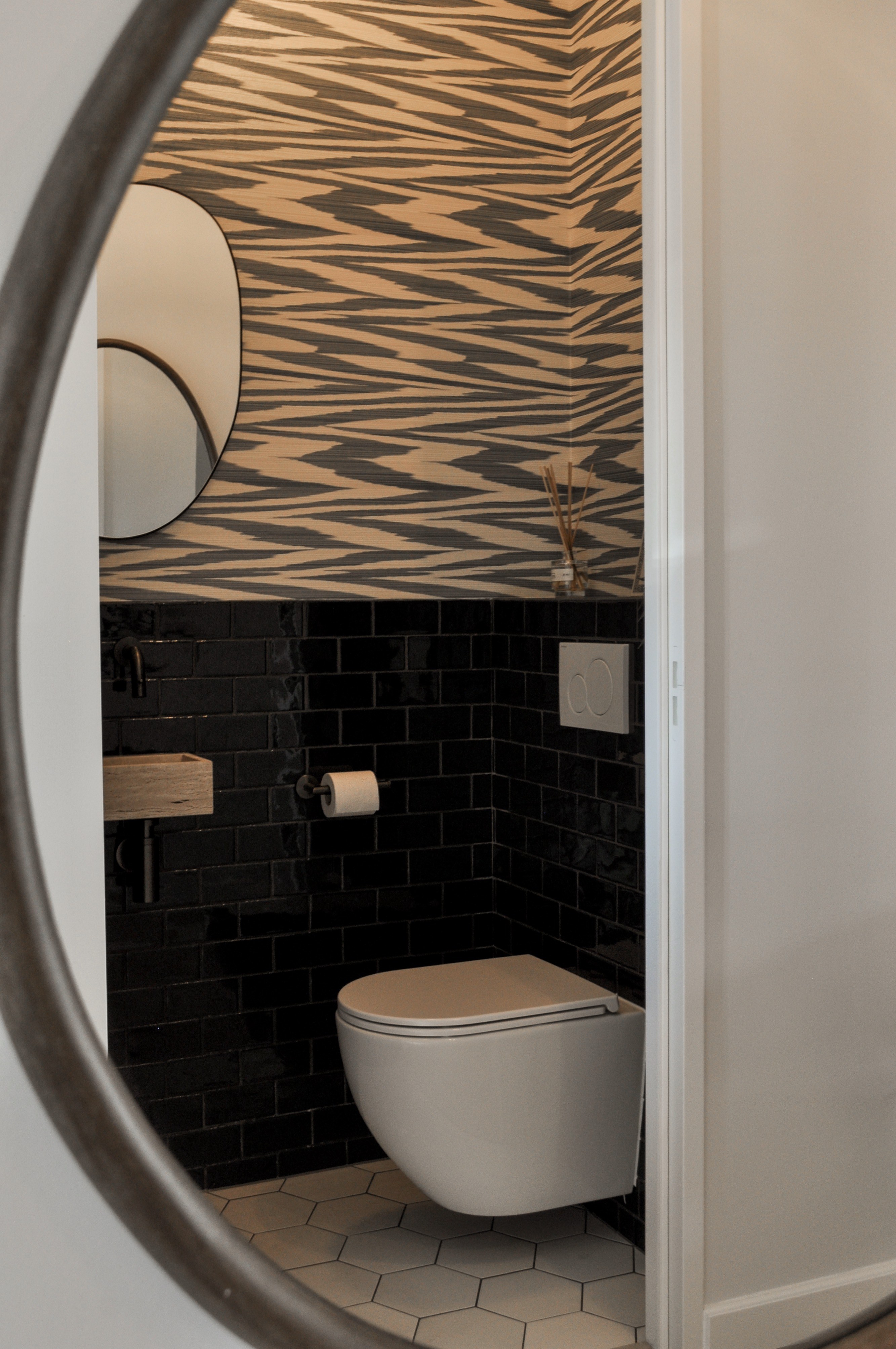 Toilet in ronde spiegel met zwarte glanzende tegels en zwart-wit grafisch behang.