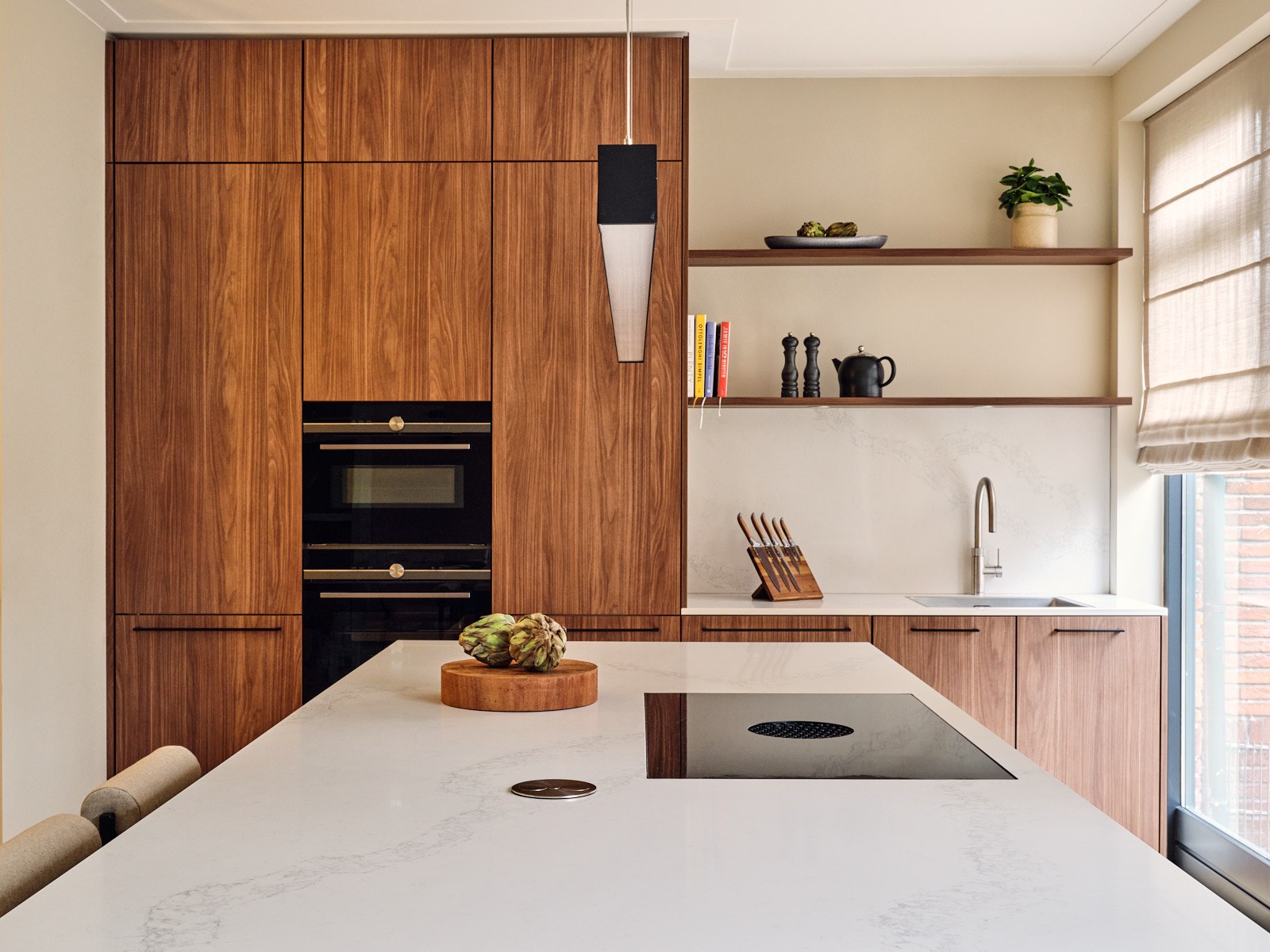 Luxe keuken in Bussum met hoge walnoot kastenwand, wit natuurstenen kookeiland, open houten wandplanken, zwarte apparatuur en minimalistische hanglamp.