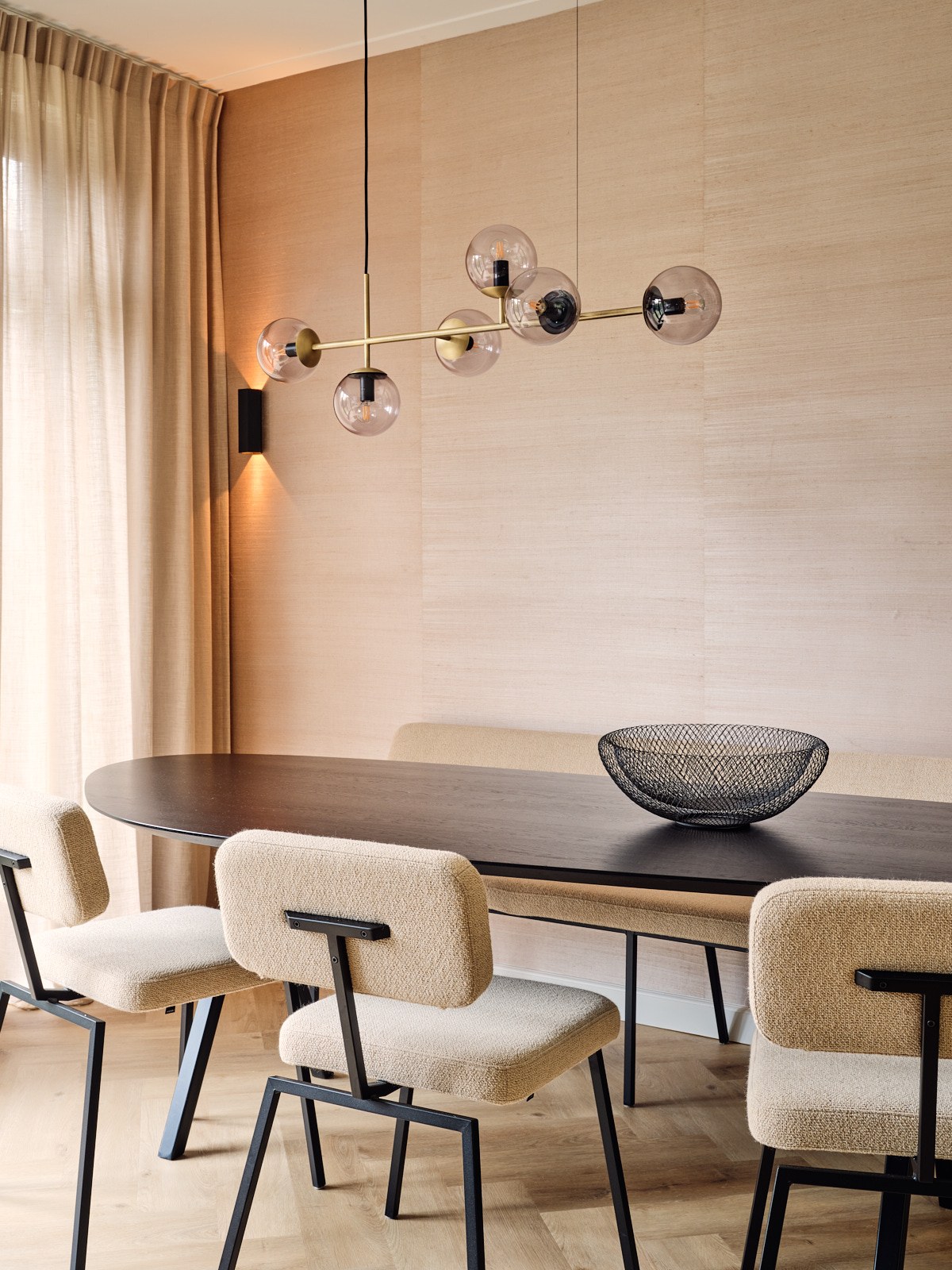 Moderne eetkamer in Bussum met donkerhouten tafel, crèmekleurige bouclé stoelen, zandkleurige houten wand, zachte gordijnen en speelse glazen verlichting.