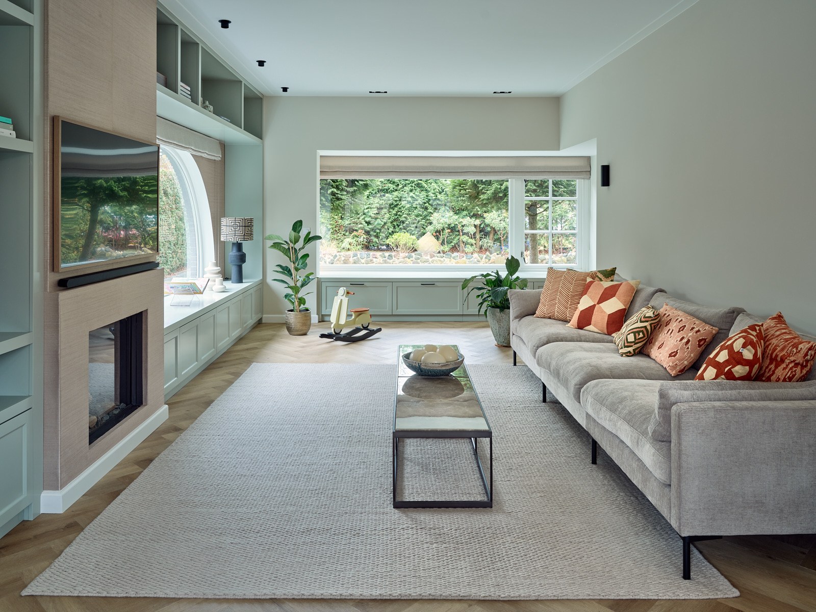 Warme moderne woonkamer in Bussum met lichtgrijze hoekbank, grijsgroene maatwerkkasten, ingebouwde haard, natuurstenen salontafel, visgraatvloer en terracotta sierkussens.