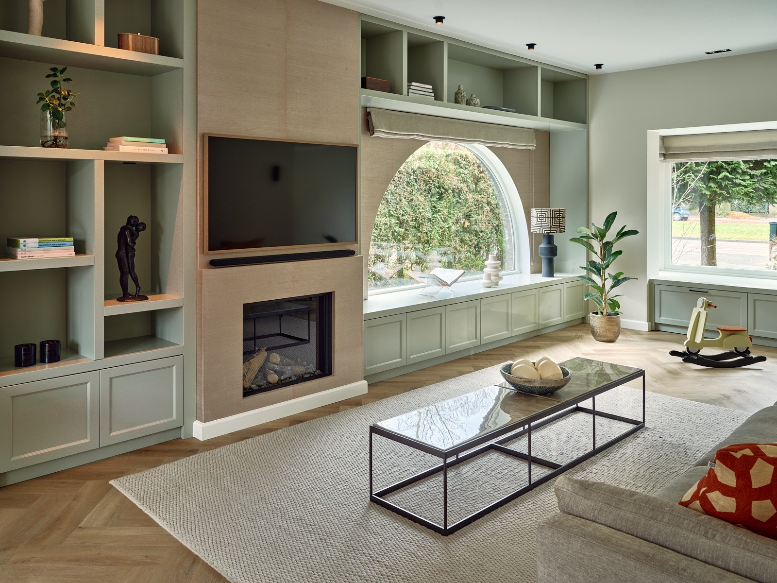 Moderne woonkamer in Bussum met grijsgroene maatwerkwand, open vakkenkast, geïntegreerde televisie en haard, lichte bank, visgraatvloer en zwart metalen salontafel.