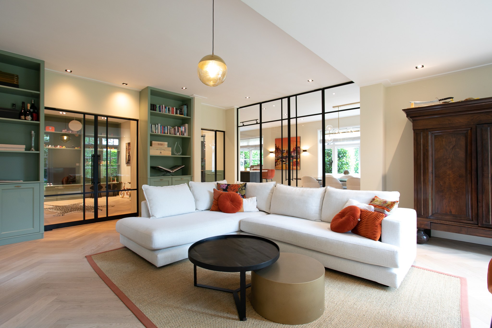 Warm moderne woonkamer met grote witte hoekbank, grijsgroene maatwerkkasten, zwarte stalen glazen wand, visgraatvloer en terracotta sierkussens.