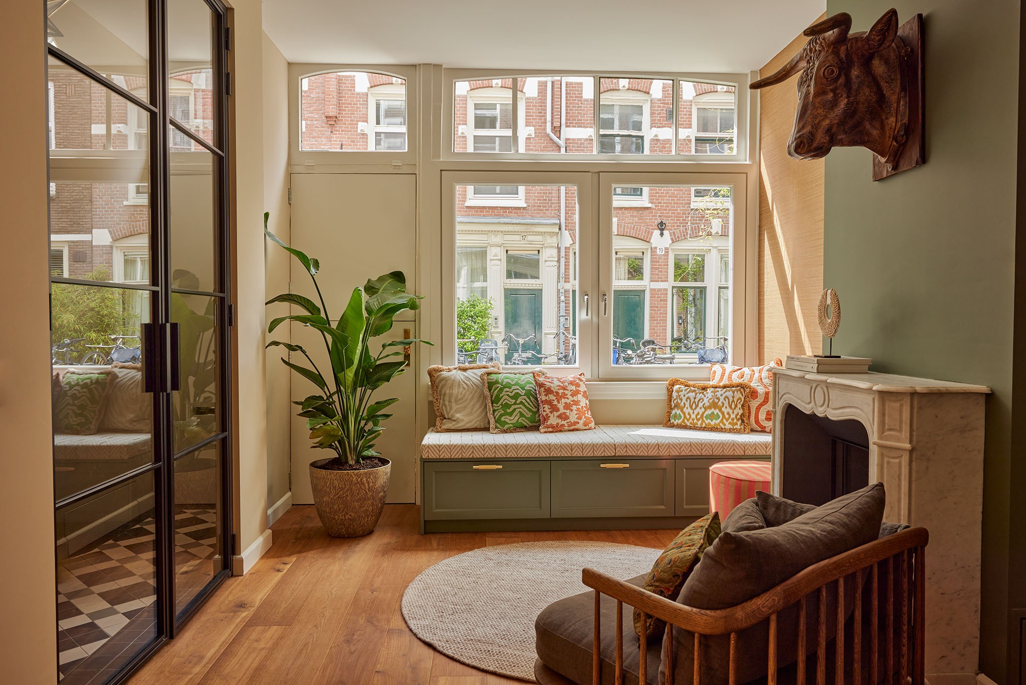 Brede woonkamer met olijfgroene wand, haard, ingebouwde raamzitbank en visgraatvloer.