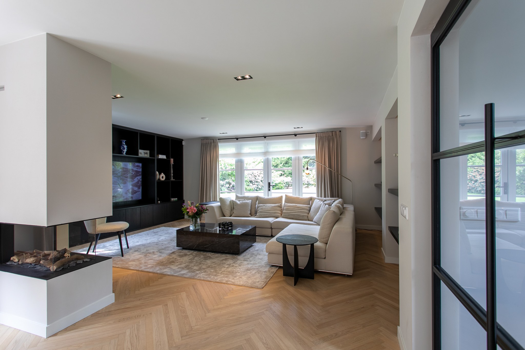 Moderne woonkamer in Blaricum met zwarte maatwerkwand rond televisie, driezijdige haard, grote beige hoekbank, natuurstenen salontafel en visgraatvloer.