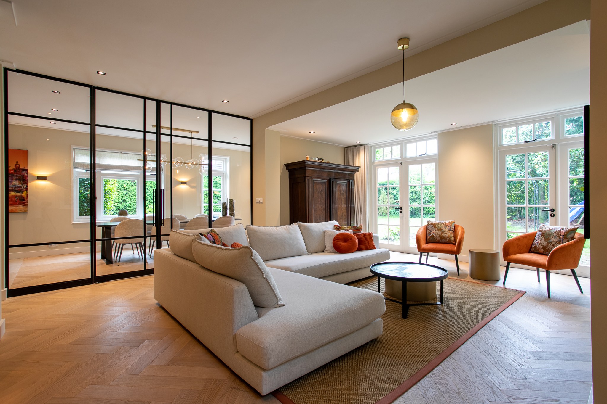 Ruime woonkamer met lichte modulaire bank, visgraatvloer, zwarte stalen glazen kamerwand, cognackleurige fauteuils en zacht terracotta textiel.