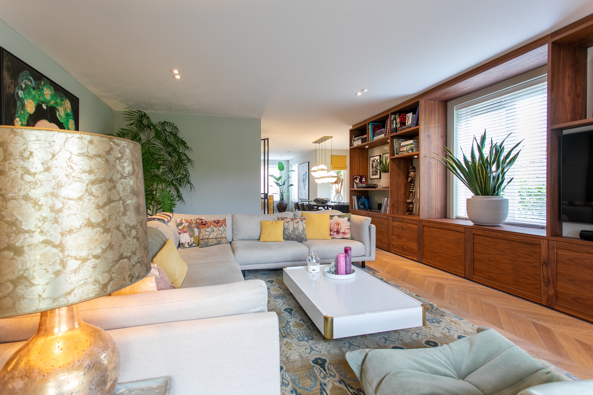 Eclectische woonkamer in Berkel en Rodenrijs met lange walnoot maatwerkwand, lichte hoekbank, visgraatvloer, grijsgroene wand en kleurrijke kunst en kussens.