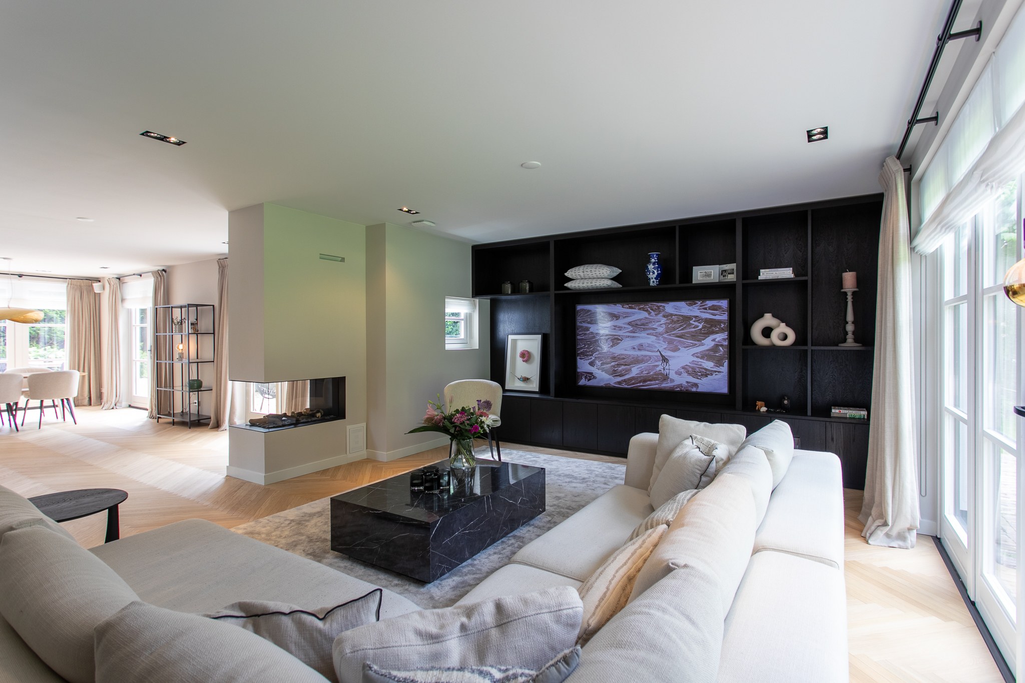 Luxe woonkamer in Blaricum met kamerbrede zwarte maatwerkkast, geïntegreerde televisie, driezijdige haard, beige hoekbank, zwarte marmeren salontafel en visgraatvloer.