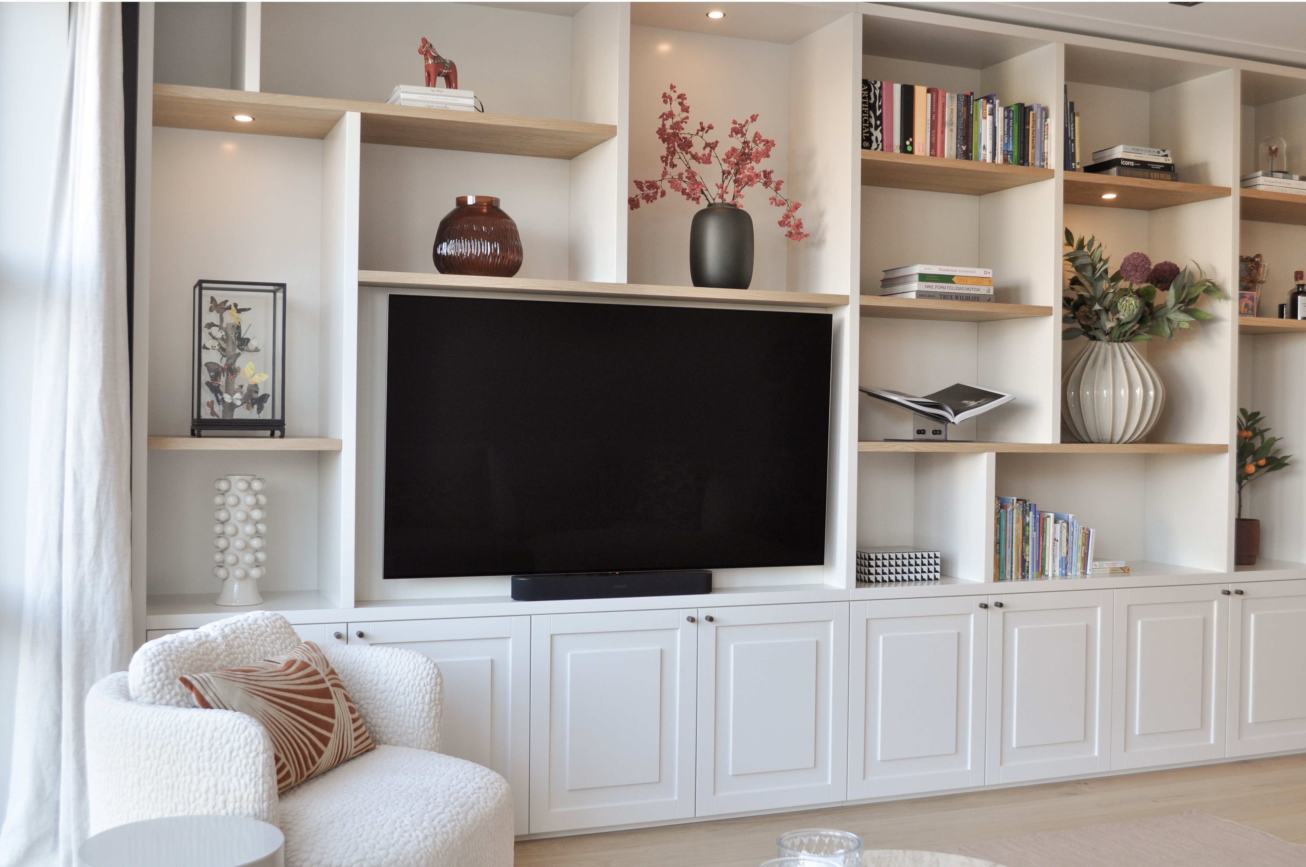 Woonkamer met witte maatwerk mediawand, geïntegreerde televisie, eiken planken en crèmekleurige fauteuil.