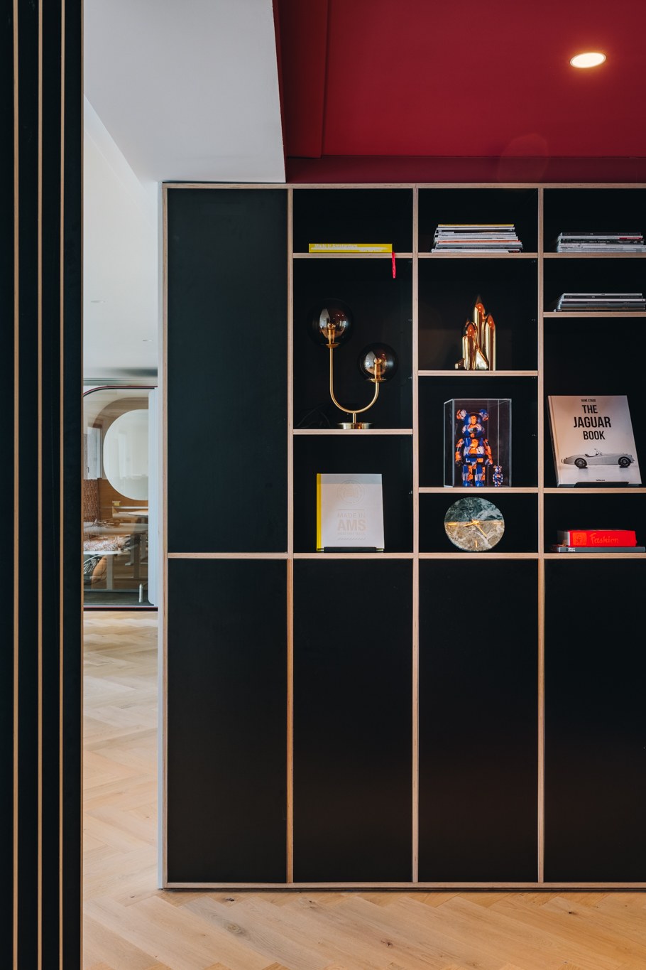 Kantoorinterieur met kamerhoge matzwarte maatwerkkast, open vakken met kunst en boeken, fijne goudkleurige belijning en bordeauxrood plafondaccent.
